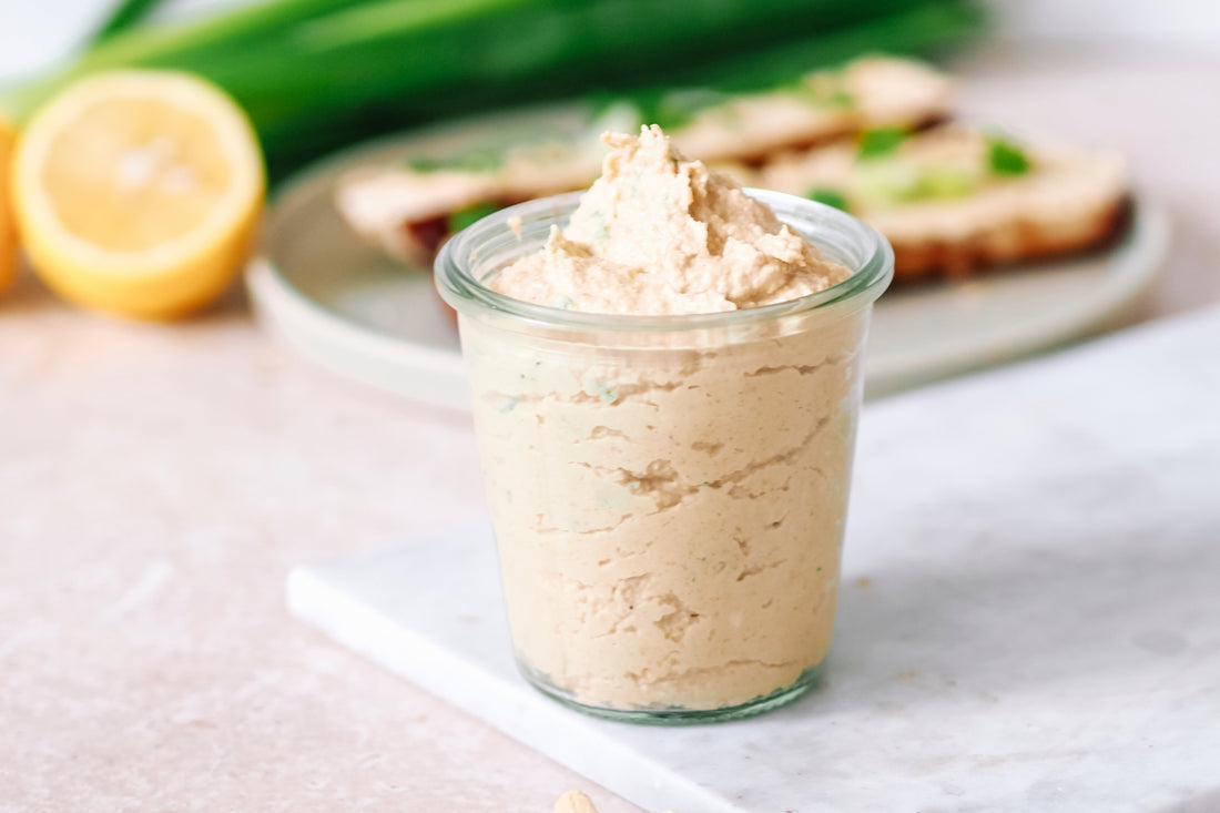 Veganer Obazda im Glas, serviert neben einer belegten Scheibe Brot mit grünen Frühlingszwiebeln und einem Hintergrund aus frischen Zutaten wie Lauch und Zitrone auf einer hellen Arbeitsplatte